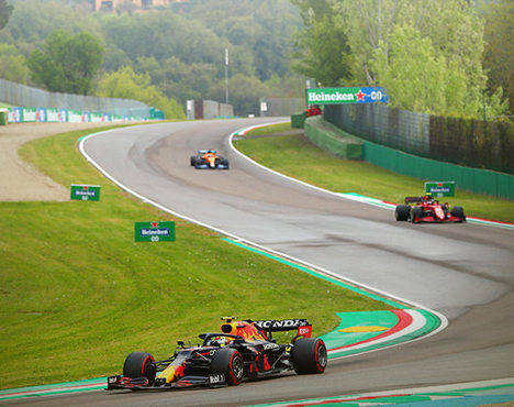 Формула-1: на гоночной трассе автомобиль Red Bull идёт впереди машины McLaren и Ferrari. Трасса извилистая с зелёными зонами за пределами асфальта. На заднем плане видна реклама Heineken и зелёные деревья