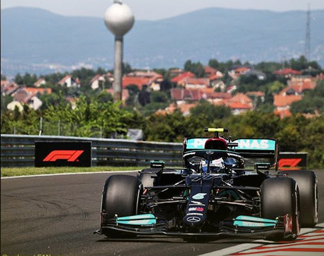 Гоночный автомобиль с логотипом Mercedes и Petronas на трассе. На фоне видны дома и водонапорная башня. Машина участвует в автогонке, огражденная рекламными баннерами Формулы-1