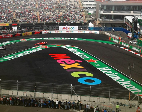 Автодром с трибунами и зрителями. На трассе видна яркая надпись Mexico и логотип Heineken. Несколько гоночных машин проходят поворот. Трасса окружена барьерами и рекламными баннерами